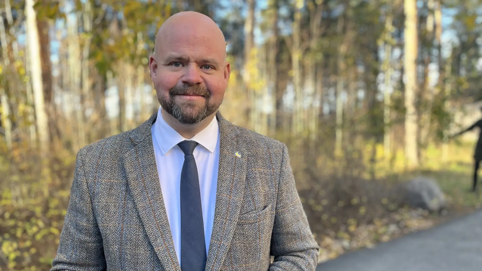 Peter Kullgren, landsbygdsministern, står utomhus i Luleå iklädd kostym och slips för att presentera upphandlingen av ett nytt beredskapslager. Bakgrunden består av träd och en asfalterad väg.