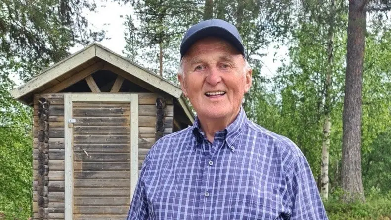 Harry, 92, står framför en timrad byggnad i en skogsmiljö. Han bär blå keps och rutig skjorta.