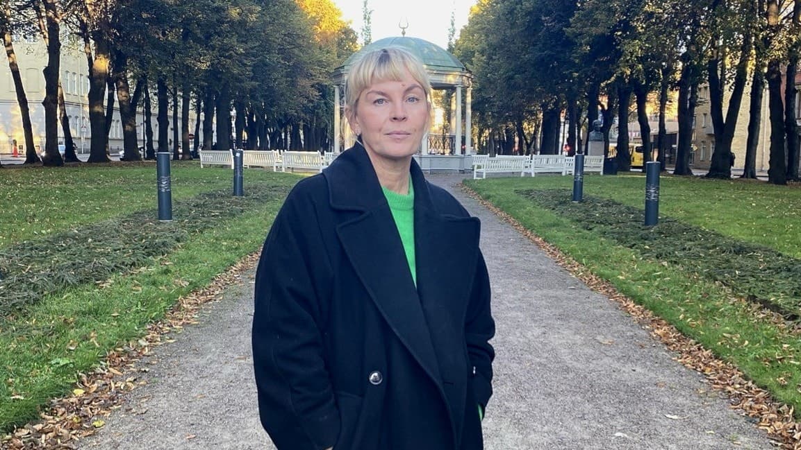 Amelie Edvinsson från Kvinnojouren Blåklockan står på en grusgång omgiven av höstklädda träd i en park. Hon bär en svart kappa och grön tröja, med stadsmiljö i bakgrunden.