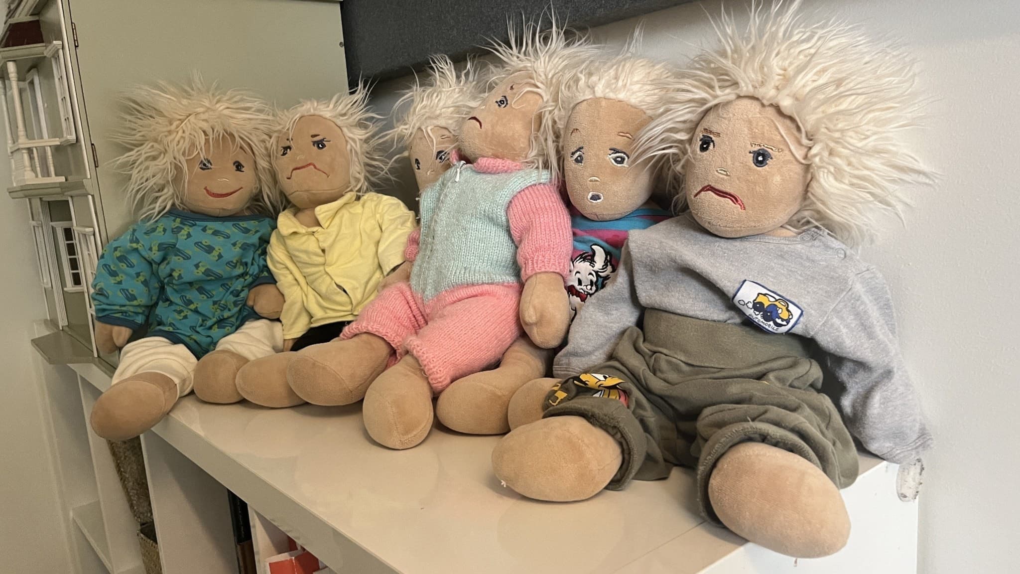 Fem mjukdockor med olika kläder sitter på en hylla, placerade i rad, på ett skyddat boende. Dockorna har glada ansikten och spretigt, ljust hår, omgivna av en neutral inomhusmiljö.