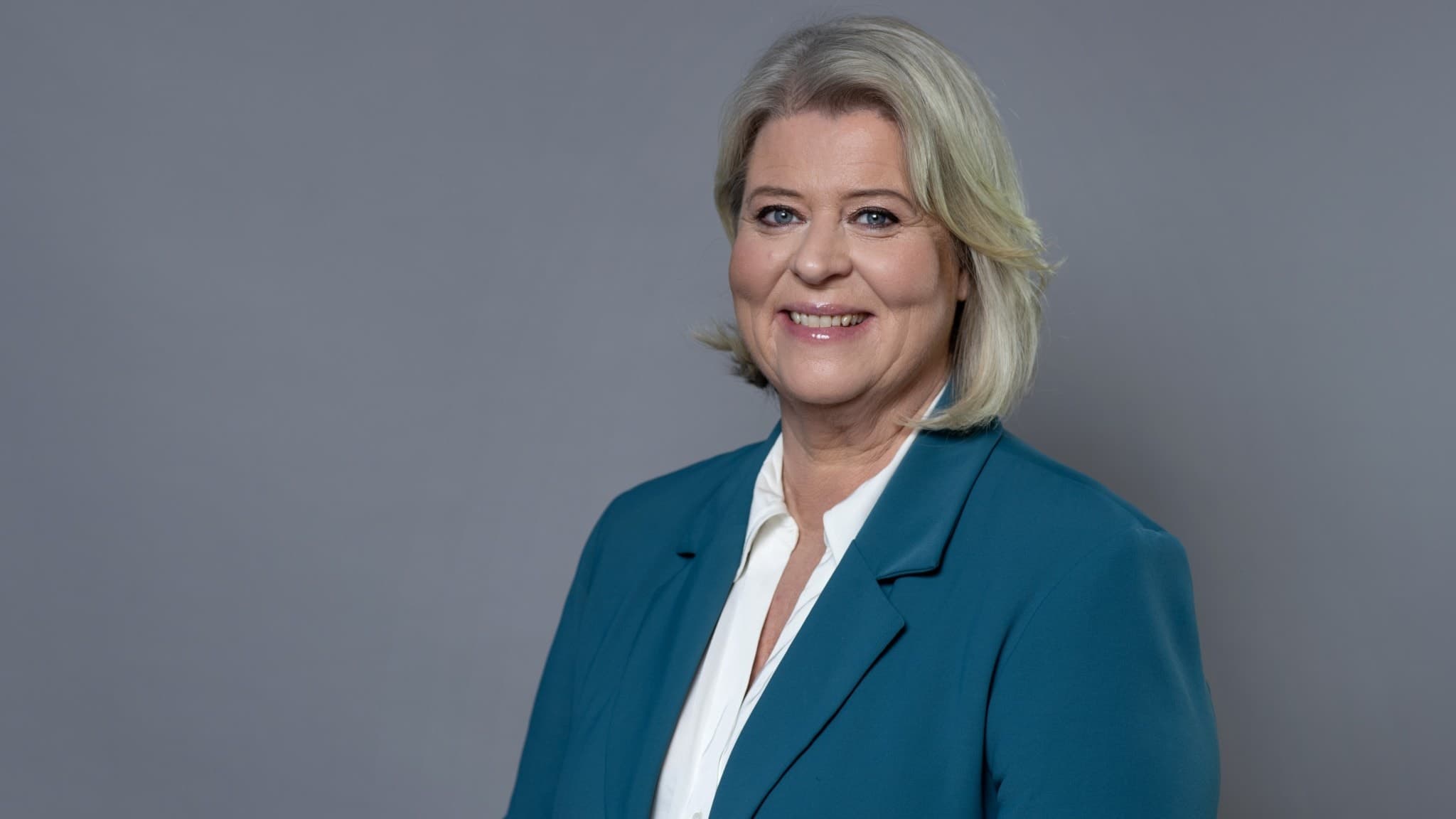 Porträttbild i halvfigur av Camilla Waltersson Grönvall, socialförsäkringsminister, klädd i en blågrön kavaj och vit blus. Hon har kort blont hår och står framför en grå bakgrund.