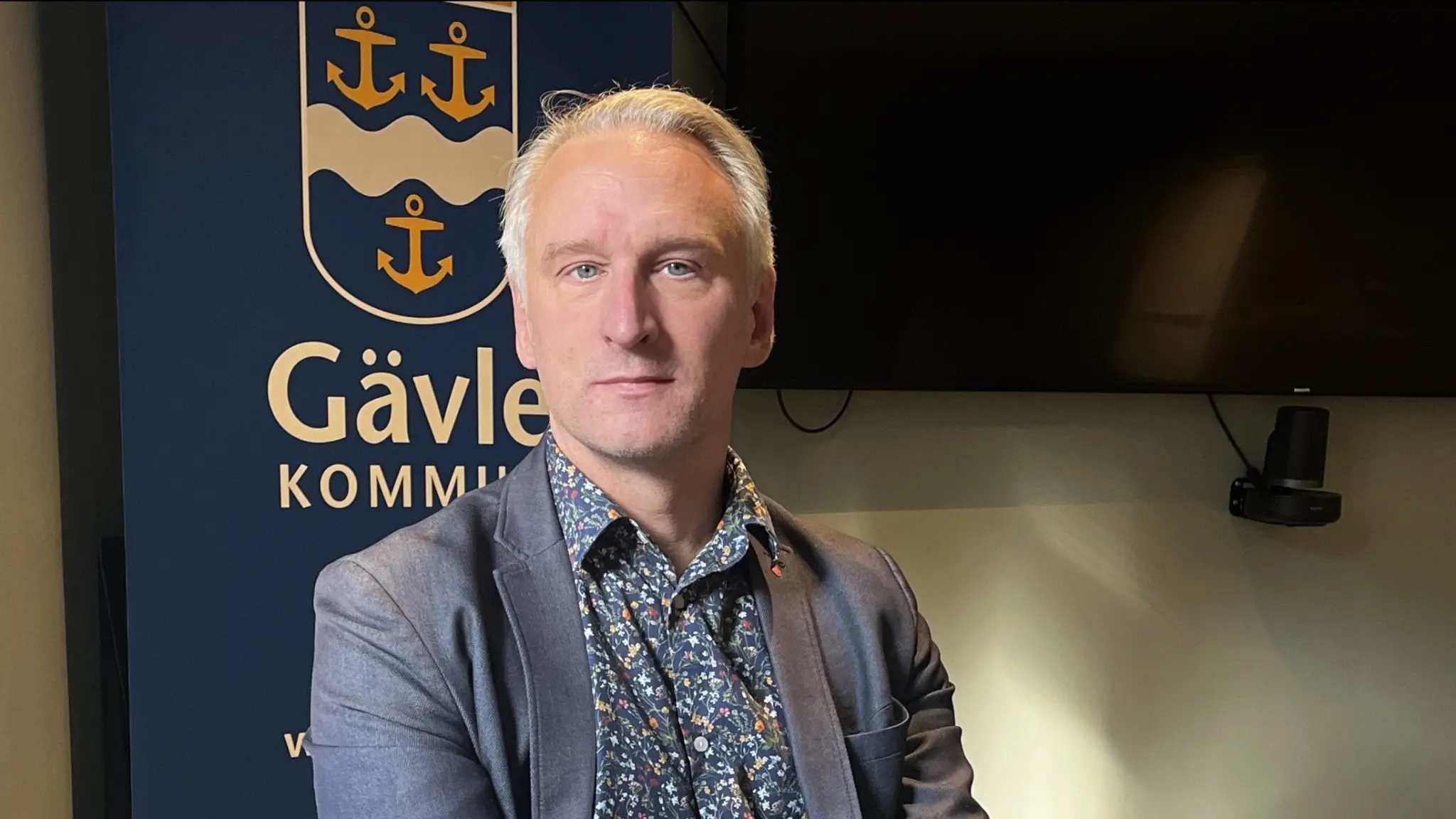 Jörgen Edsvik, kommunalråd för Socialdemokraterna i Gävle, sitter i ett rum med Gävle kommuns logotyp synlig i bakgrunden. Han bär en kavaj och skjorta med mönster.