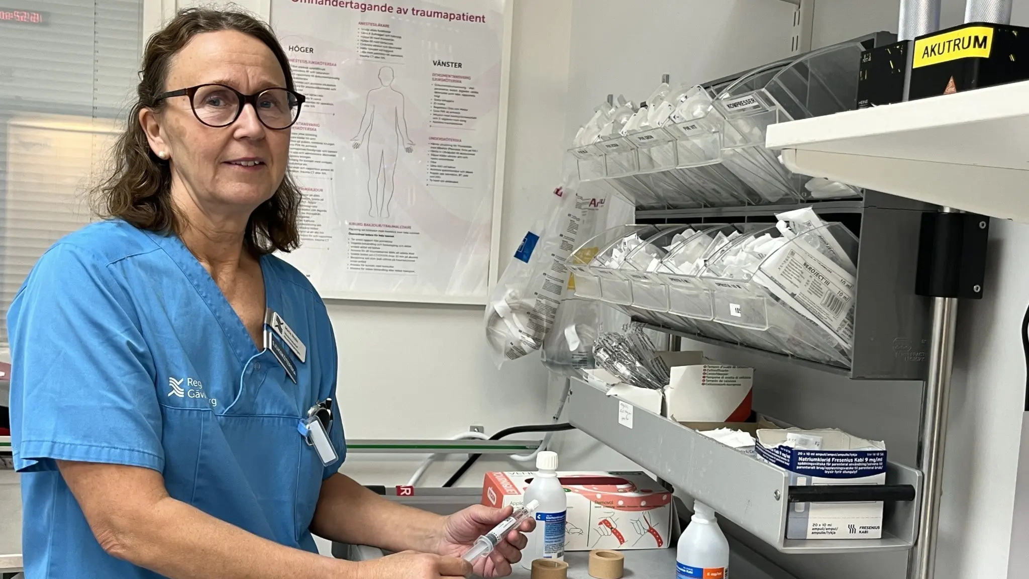 Marie Molin, vårdenhetschef vid Gävle sjukhus, står i en medicinsk miljö och håller i medicinsk utrustning med hyllor fyllda av förbandsmaterial och sjukvårdsartiklar i bakgrunden.