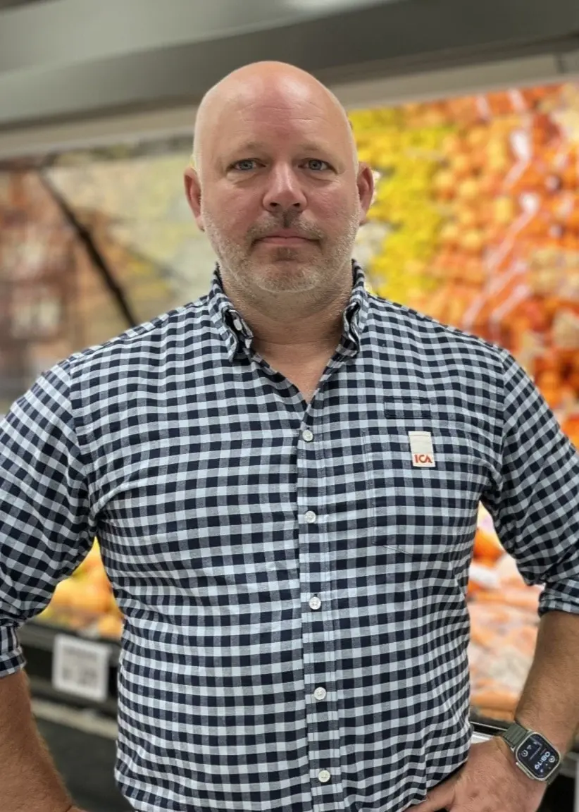 En man i rutig skjorta står framför frukt- och grönsaksavdelningen i en ICA-butik. Hyllorna är fyllda med färska grönsaker och frukter, inklusive äpplen, citrusfrukter och kål.