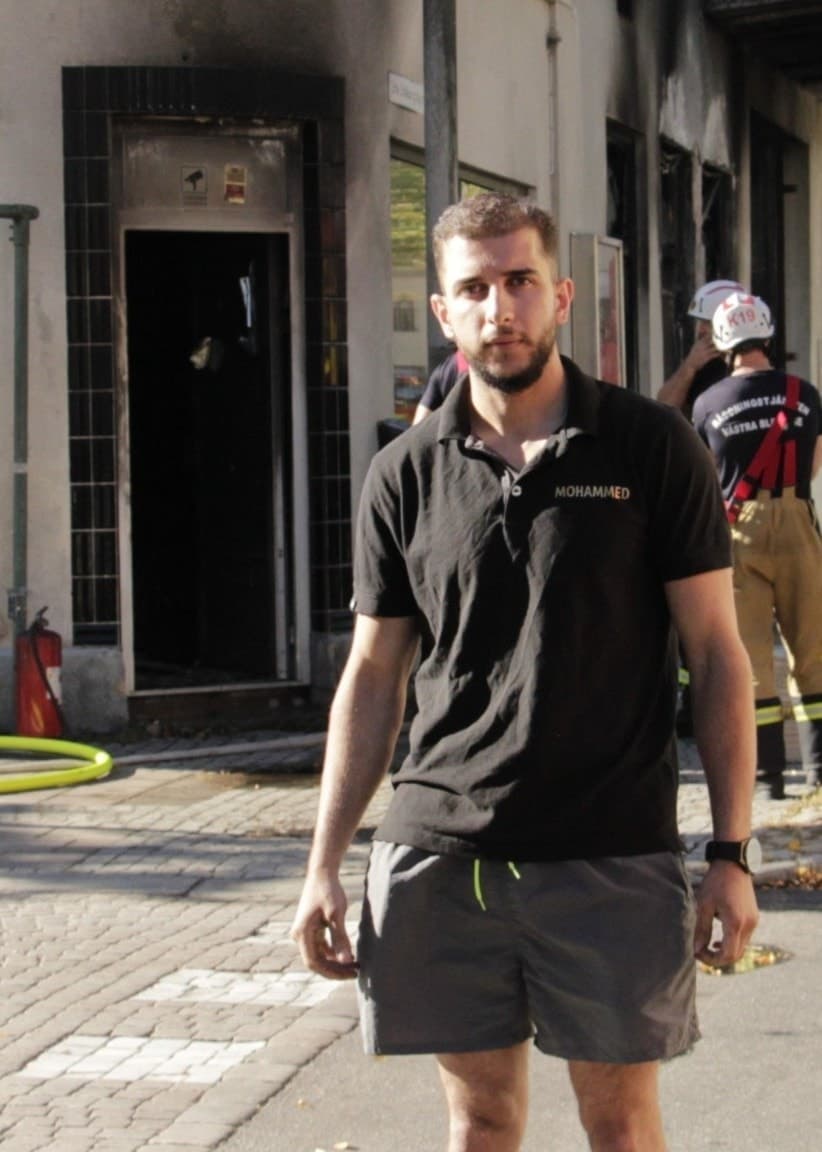 Mohammed Aldahdooh står på gatan utanför sin utbrända cykelbutik. Bakom honom syns brandmän och brandbilar.