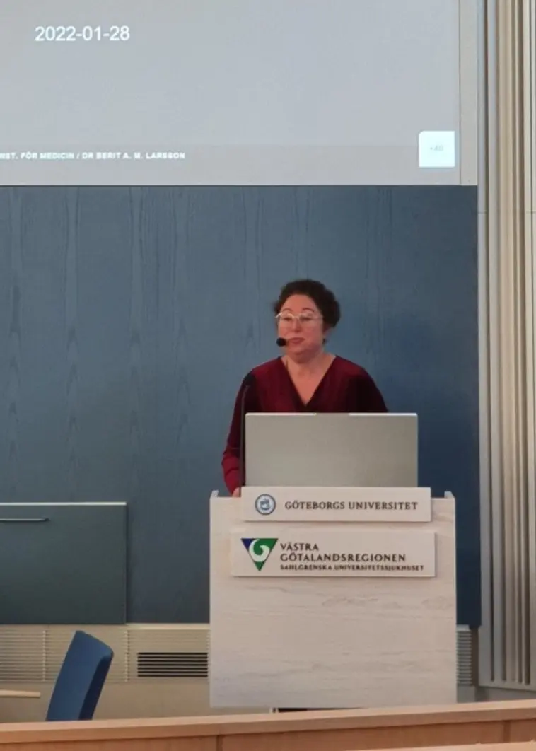Berit Larsson står vid ett podie, med mick och en presentation uppe på en digital skärm i bakgrunden,  inuti en byggnad vid Göteborgs Universitet. 