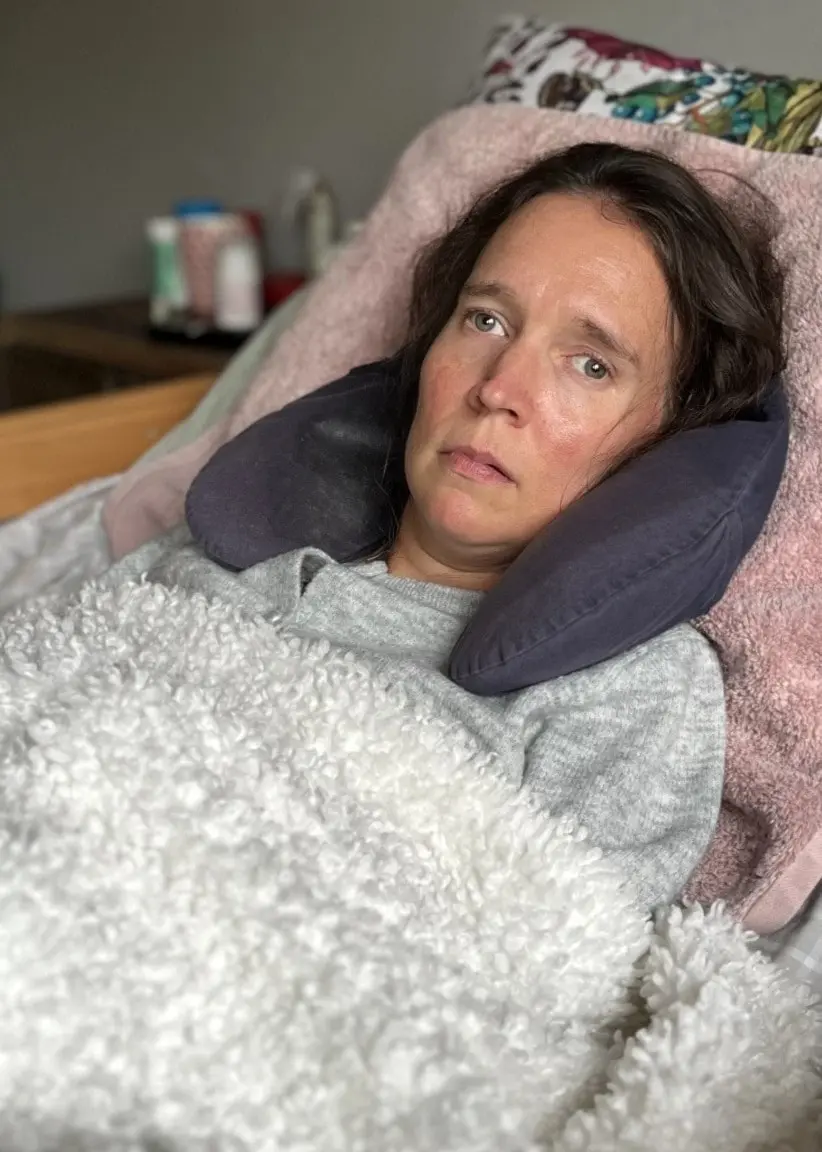 En person ligger i en säng med en ljusgrå filt över sig och huvudet vilande på en kudde. I bakgrunden syns en del av ett kök och en stol.