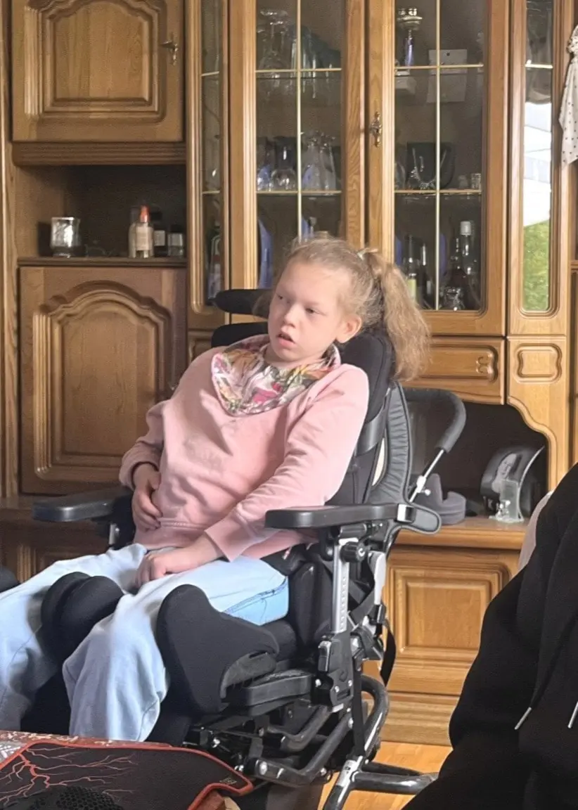Nelly sitter i en rullstol bredvid mamma Pernilla iklädd svart tröja. Bakgrunden visar ett träskåp och växter i ett vardagsrum med dämpad belysning.