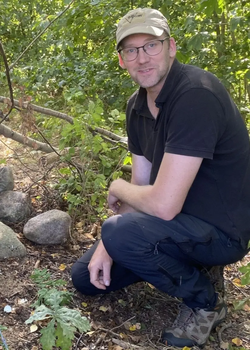 Victor Karlsson, yrkesjägare i Skåne, hukar vid en rörliknande konstruktion omgiven av träd och stenar, som används för att bekämpa invasiva arter i naturen.