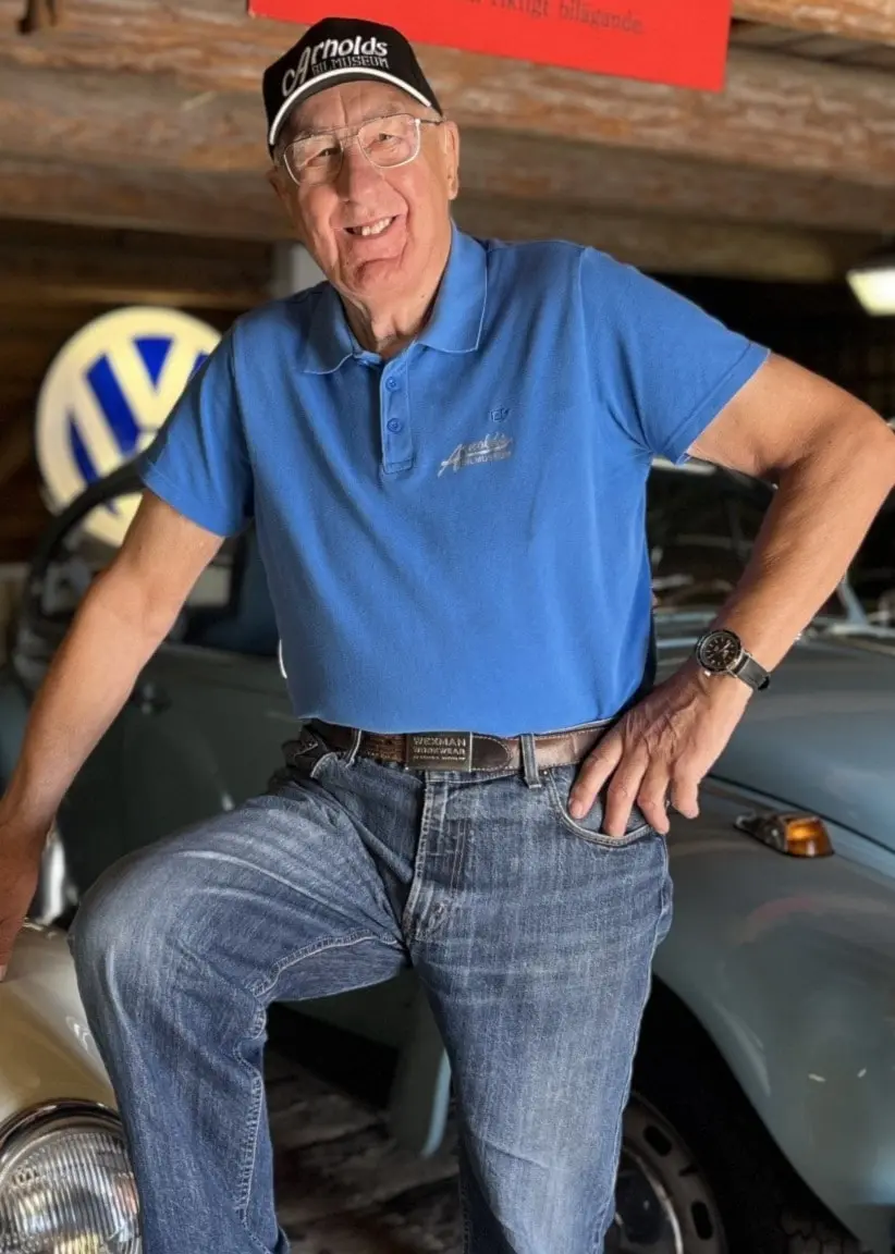Arnold Sundström poserar bredvid en vit veteranbil i ett museum med träinteriör, omgiven av ytterligare klassiska bilar och nostalgiska skyltar.