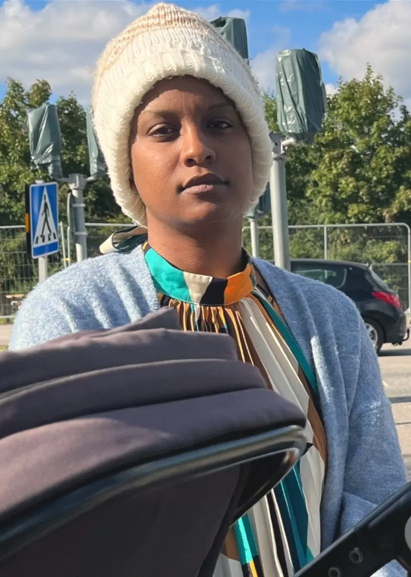 Ariam sitter med sin dotter i barnvagn vid ett övergångsställe i Gottsunda. I bakgrunden syns träd, höghus och en bil.