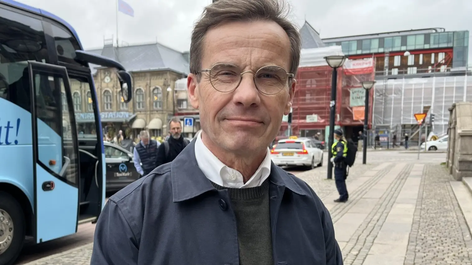 En person står framför en blå buss med texten 'Framåt' på sidan, på en trottoar i stadsmiljö. I bakgrunden syns byggnader, folk och en mulen himmel.