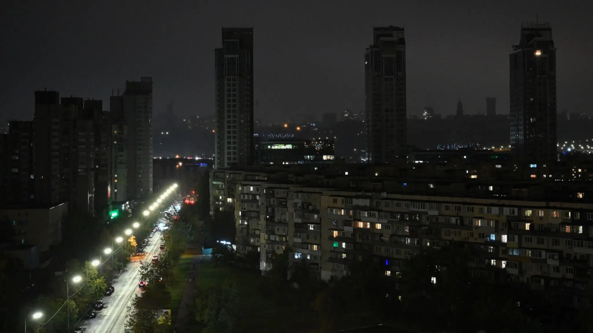 Mörklagda bostadshus vid Dniproflodens östra strand i Kyiv under ett strömavbrott orsakat av ryska missil- och drönarattacker, med en svagt upplyst gata i förgrunden.